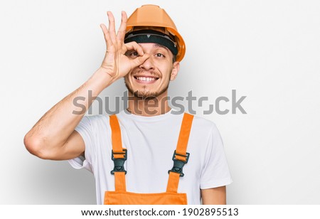 Hispanic young man wearing handyman uniform and safety hardhat smiling happy doing ok sign with hand on eye looking through fingers 