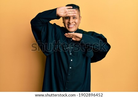 Young latin priest man standing over yellow background smiling cheerful playing peek a boo with hands showing face. surprised and exited 