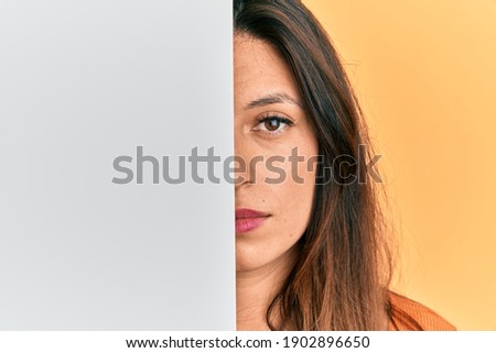 Beautiful hispanic woman holding blank empty banner covering half face relaxed with serious expression on face. simple and natural looking at the camera. 