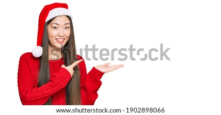 Young chinese woman wearing christmas hat amazed and smiling to the camera while presenting with hand and pointing with finger. 