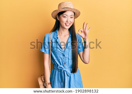 Young chinese woman wearing summer hat smiling positive doing ok sign with hand and fingers. successful expression. 