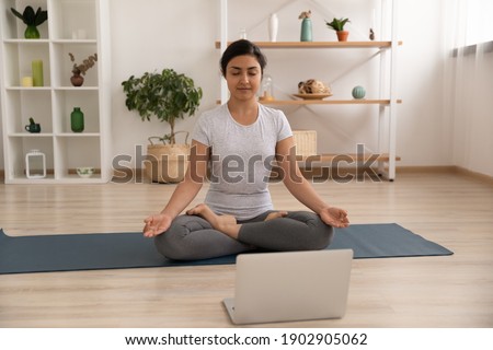 Concentrated indian woman sit on gymnastic mat in lotus pose before laptop screen meditate listen to calm music. Peaceful young hindu lady practice yoga at home learn asanas from online training class Royalty-Free Stock Photo #1902905062