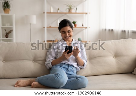 Happy indian lady relax at home alone sit on large couch in comfortable pose share good news at social media via cellphone. Smiling mixed race woman enjoy weekend order goods food online in phone app Royalty-Free Stock Photo #1902906148