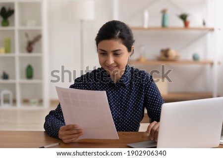 Confident indian female small business owner sit by desk with laptop study paper letter of supplier choose samples of production to order buy. Young hindu woman freelancer doing paperwork at workplace Royalty-Free Stock Photo #1902906340