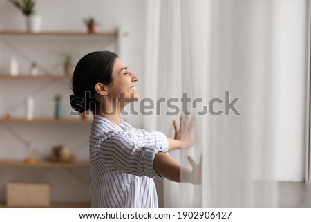 Bathing in sunlight. Excited young indian lady meet first morning at new flat house part curtains enjoy being homeowner. Happy hindu female open drapes on window breath fresh air close eyes in delight Royalty-Free Stock Photo #1902906427