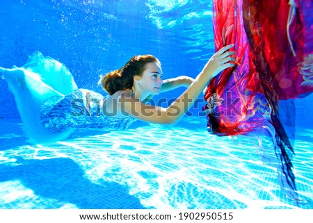 A young girl in a shiny blue dress swims underwater in a pool and catches a red cloth that sinks in the water. Portrait. Side view. Underwater photography. Horizontal orientation.