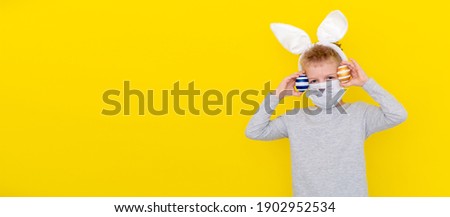 Banner copyspace Boy in rabbit bunny ears on head and protective mask with colored eggs on yellow studio background. Cheerful crazy smiling happy child. Covid easter kid.