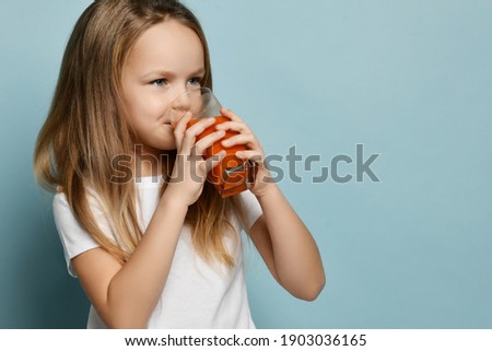 Portrait of happy smiling cute kid girl in white t-shirt drinking enjoying fresh fruit juice and looking at copy space over blue background. Happy childhood, healthy lifestyle concept