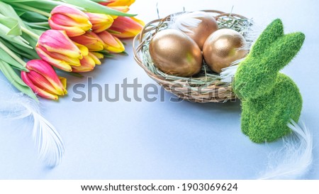 Easter eggs basket. Golden egg in basket with spring tulips, white feathers on pastel blue background in Happy Easter decoration. Traditional decoration in sun light