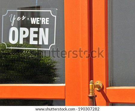 Cordial Invitation: Vibrant 'Yes, We Are Open' Sign Hung on an Eye-Catching Orange Wooden Door with Transparent Glass Pane