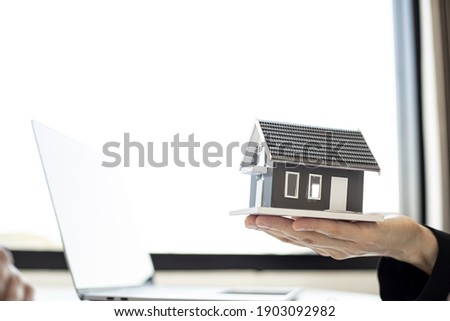 A female home salesman holds a gray miniature house model to describe the details of the house to the customer who will purchase a house on the project. Real estate trading ideas.