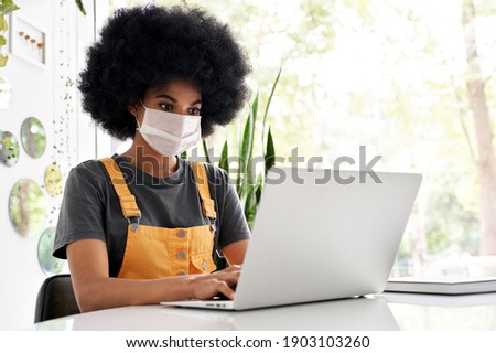 Young African American teen girl student with Afro hair wearing face mask using laptop sitting at cozy cafe table alone indoor. Social distancing and work, study on computer with covid 19 protection. Royalty-Free Stock Photo #1903103260