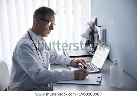 Serious mature old doctor physician making medical notes in notebook using laptop in hospital office. Senior middle aged male practitioner working on computer and writing sitting at desk. Royalty-Free Stock Photo #1903103989