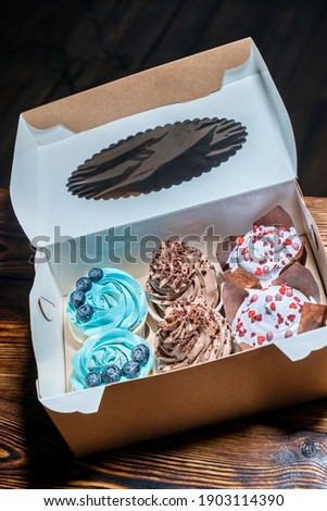 Delivery in carton box of cupcakes or fairy cakes decorated with blueberries and blue creamy-cheese, brown cocoa topping and white cream on dark wooden table. Vertical orientation image