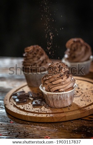 Side view of three sweet homemade cupcakes or fairy cakes decorated with brown cocoa creamy cheese topping and falling chocolate chips on wooden cutting board on table. Vertical image, selective focus