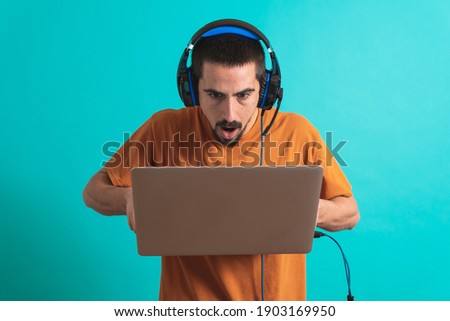 Young handsome man with gamer headphone set isolated over blue background using laptop computer playing a game. Professional gamer playing video game on his personal computer.