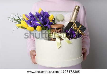 Woman holding basket with gifts, bouquet and champagne on light grey background, closeup Royalty-Free Stock Photo #1903172305