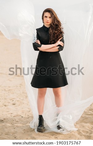 Confident woman. Female rights. Independence freedom. Concerned brunette lady in black casual dress standing with folded arms in sandy desert with flying polyethylene film.
