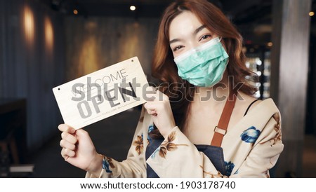 Business owner attractive young Asian woman in apron hanging we're open sign on front door  welcoming clients to new cafe. Happy waiter with protective face mask holding open sign at cafe .