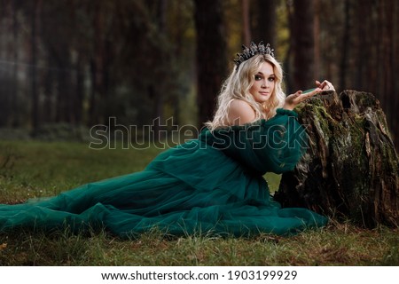 A beautiful blonde young woman in a long green dress and a diadem on her head in the forest. girl sitting near the old stump. Solar glare. Fantasy. fairy tale.