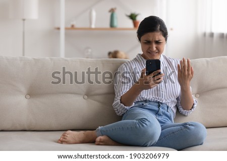 Confused angry indian woman sit on couch at home read spam message on phone screen have problem with correct work. Shocked mad mixed race lady web store client deal with scam online. Copy space Royalty-Free Stock Photo #1903206979
