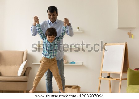 Overjoyed millennial indian dad and little kid boy having fun together play funny active game at home. Laughing young mixed race father holding small son hands allowing him jump high raising up to air Royalty-Free Stock Photo #1903207066