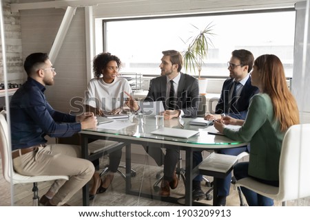 Interested diverse colleagues listening to leader, discussing statistics, sitting at table in modern boardroom, mentor coach explaining strategy, giving instructions to workers at briefing Royalty-Free Stock Photo #1903209913