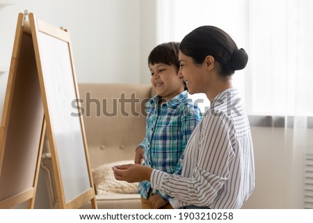Friendly indian woman psychologist and small boy stand by easel board hold crayon planning picture talk on visual art therapy session. Happy hindu mother teach little son to draw using piece of chalk