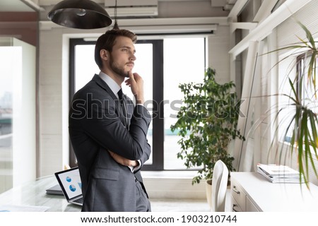 Thoughtful businessman wearing suit touching chin, looking to aside, standing in modern office, executive manager leader pondering future or project strategy, planning workday, lost in thoughts Royalty-Free Stock Photo #1903210744