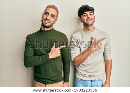Homosexual gay couple standing together wearing casual clothes smiling cheerful pointing with hand and finger up to the side 
