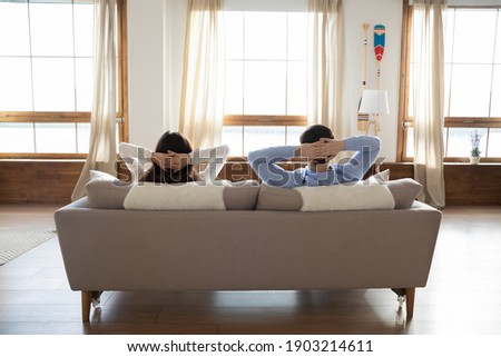 Rear view full length young couple relaxing on couch in modern apartment, young woman and man leaning back with hands behind head, resting, enjoying lazy weekend at home, moving day or mortgage Royalty-Free Stock Photo #1903214611