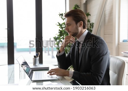 Side view thoughtful businessman looking at laptop screen, pondering task, online project strategy or financial report, pensive entrepreneur touching chin, using computer, sitting at desk in office