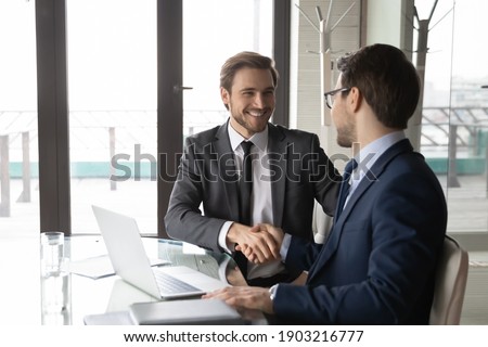 Smiling businessman shaking client hand, closing successful deal, sitting at table with laptop in office, satisfied hr manager hiring new employee, business partners handshaking at meeting Royalty-Free Stock Photo #1903216777