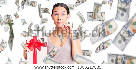 Beautiful brunette young woman holding gift serious face thinking about question with hand on chin, thoughtful about confusing idea