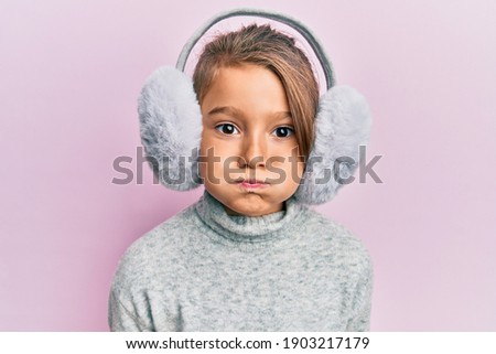 Little beautiful girl wearing fluffy earmuff puffing cheeks with funny face. mouth inflated with air, catching air.  Royalty-Free Stock Photo #1903217179
