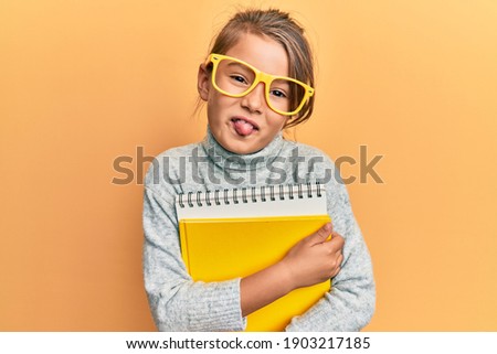 Little beautiful girl wearing glasses and holding books sticking tongue out happy with funny expression. 