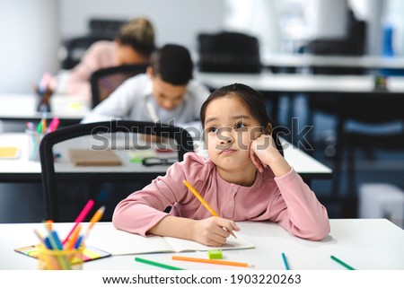 Education And Learning Concept. Portrait of tired and bored small asian girl sitting at desk in classroom at school, writing in notebook and thinking, looking away at window, resting head on hand