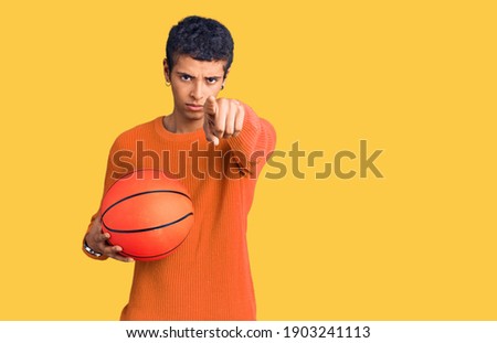 Young african amercian man holding basketball ball pointing with finger to the camera and to you, confident gesture looking serious 