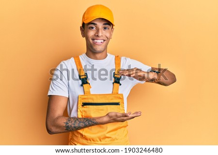 Young handsome african american man wearing handyman uniform over yellow background gesturing with hands showing big and large size sign, measure symbol. smiling looking at the camera. measuring
