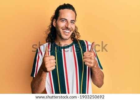 Young handsome man with long hair wearing casual clothes success sign doing positive gesture with hand, thumbs up smiling and happy. cheerful expression and winner gesture. 
