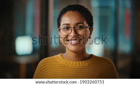 Close Up Portrait of a Young Latina with Short Dark Hair and Glasses Posing for Camera in Creative Office. Beautiful Diverse Multiethnic Hispanic Female Wearing Yellow Jumper is Happy and Smiling.