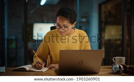 Young Hispanic Marketing Specialist Working on Laptop Computer in Busy Creative Office Environment. Beautiful Diverse Multiethnic Female Project Manager is Writing Down Notes in Paper Notebook. Royalty-Free Stock Photo #1903260901