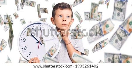Cute blond kid holding big clock serious face thinking about question with hand on chin, thoughtful about confusing idea