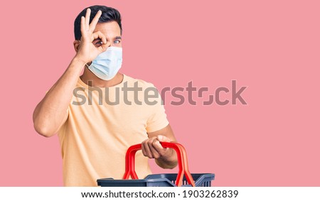 Young hispanic man wearing shopping basket and medical mask smiling happy doing ok sign with hand on eye looking through fingers 