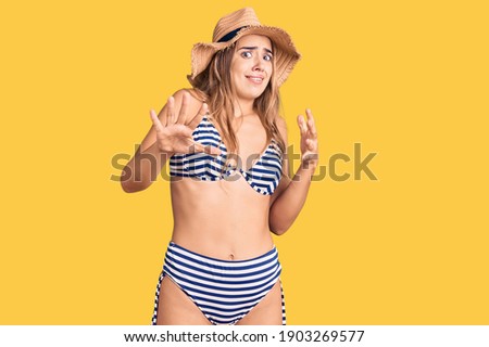 Young beautiful blonde woman wearing bikini and hat afraid and terrified with fear expression stop gesture with hands, shouting in shock. panic concept. 