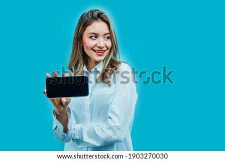Close up photo of an attractive girl on a blue background in the studio, she is holding a phone with a black screen. Online shopping. Internet. Photo for advertising.