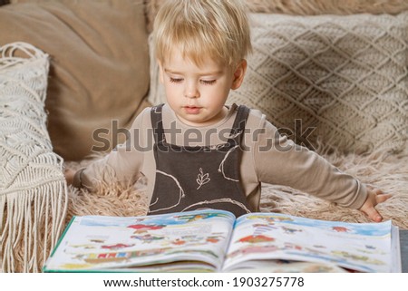Little cute blond Caucasian boy, a child reads a book with pictures, earlier development, classes with children, children at home in quarantine, the kid smiles.
