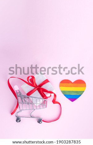 Buying LGBT gift for Valentine's Day concept. Gift box with red ribbon in shopping trolley and painted paper LGBT heart shape on pink background, copy space, flat lay, virtual frame