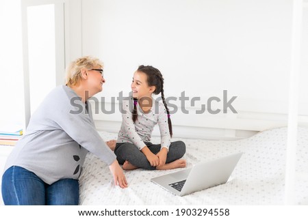 Mature grandmother helping child with homework at home. Satisfied old grandma helping her granddaughter studying in living room. Little girl writing on notebook with senior teacher sitting next to her
