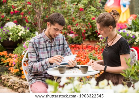 A young couple in the garden, reading books. Blossoming colorful flowers, smiling faces, relaxed summer leisure in park. Nice aromas, romantic mood. Happy boy and girl.
